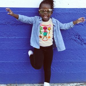 Black girl child, age 5–6, wearing a natural colour T-shirt with Mr Cutesy Myllo design, smiling and playful